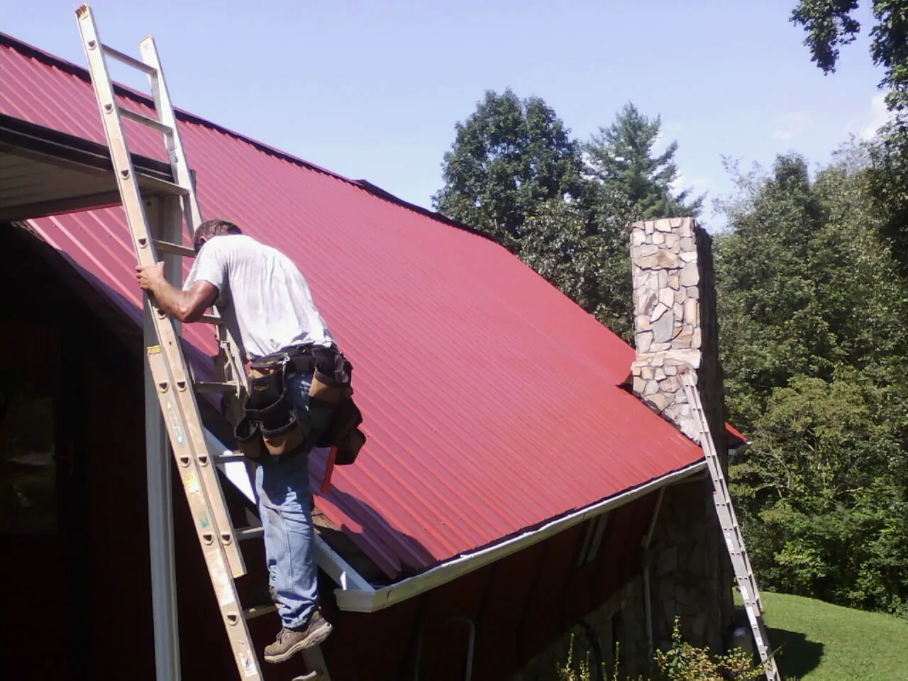 Roofer on a ladder beside a red metal roof and stone chimney — Soffit & Fascia Repair by Roofing Brothers in Saltsburg